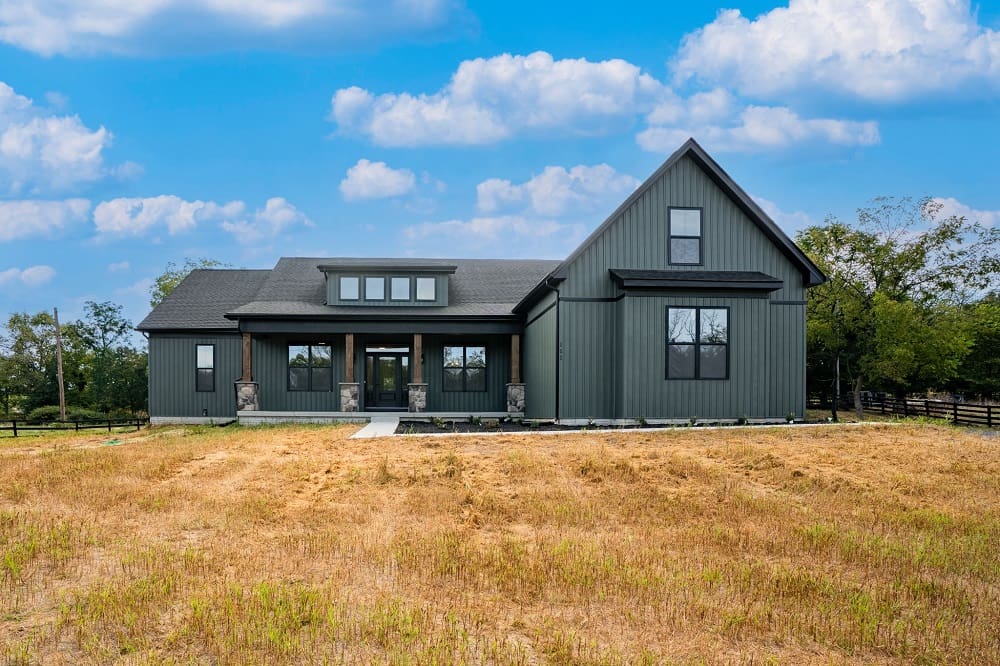 A modern dark gray farmhouse with a pitched roof stands alone in a dry golden field under a bright blue sky with scattered clouds.