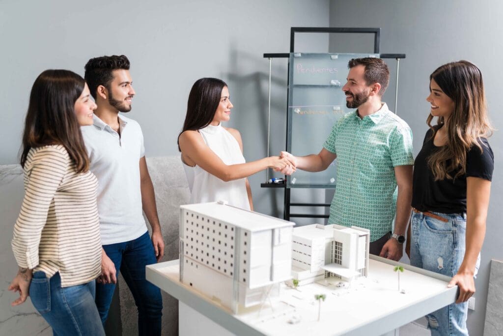 A team of five professionals collaborate around an architectural model in a modern workshop or office setting, actively discussing and presenting their design work.